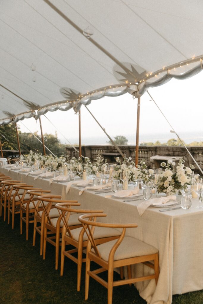 Elegant wedding tablescape under tent with neutral linens, wooden chairs, and white floral centerpieces overlooking the Crane Estate grounds.