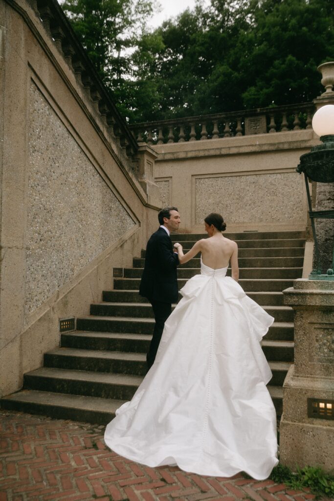 Bride in elegant gown and groom in classic tuxedo walking up stone staircase at the Crane Estate garden.