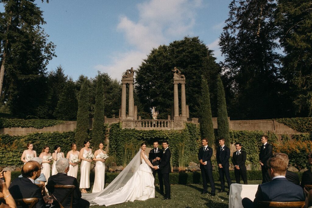Bride and groom exchange vows in a garden ceremony at the Crane Estate in Ipswich, MA, surrounded by wedding party and historic architecture.