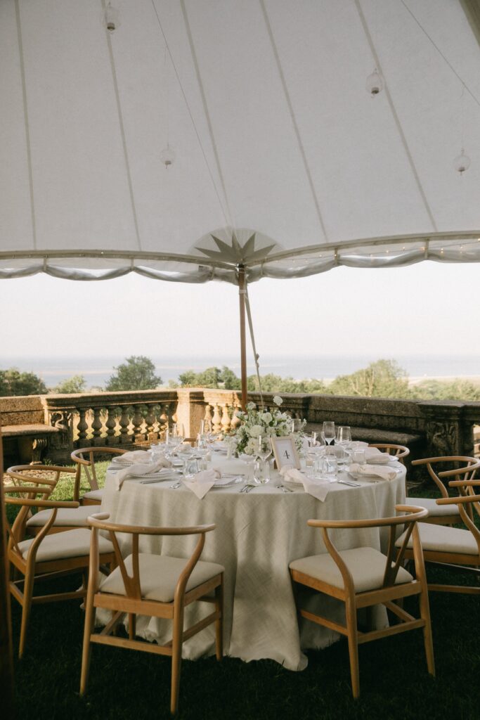 Wedding tablescape under tent with neutral linens, wooden chairs, and white florals at the Crane Estate.