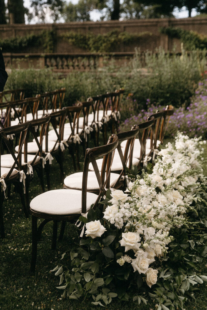 Elegant garden wedding ceremony seating with wooden cross-back chairs and lush white floral aisle arrangements at the Crane Estate.