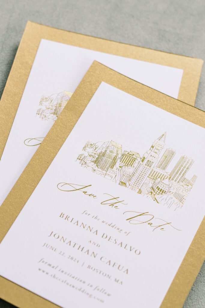 A luxe save the date card printed with metallic gold foil, featuring a custom line drawing of the Boston skyline. Mounted on shimmery gold cardstock, this suite combines elegant script with modern city-inspired design—perfect for an upscale urban wedding in Boston, Massachusetts.