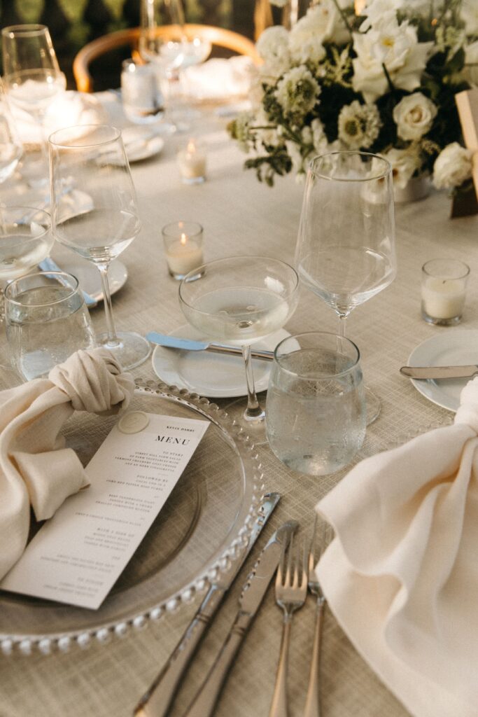 Wedding menu with vellum place card and white wax seal displayed on neutral reception table with glass plate and taupe napkin.
