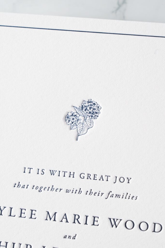 An elegant letterpress wedding invitation featuring deep navy ink pressed into soft cotton paper. A fine-line floral illustration adds a romantic touch to the top, while serif and italic typefaces offer timeless charm. This suite is ideal for couples who love traditional design with a touch of detail.