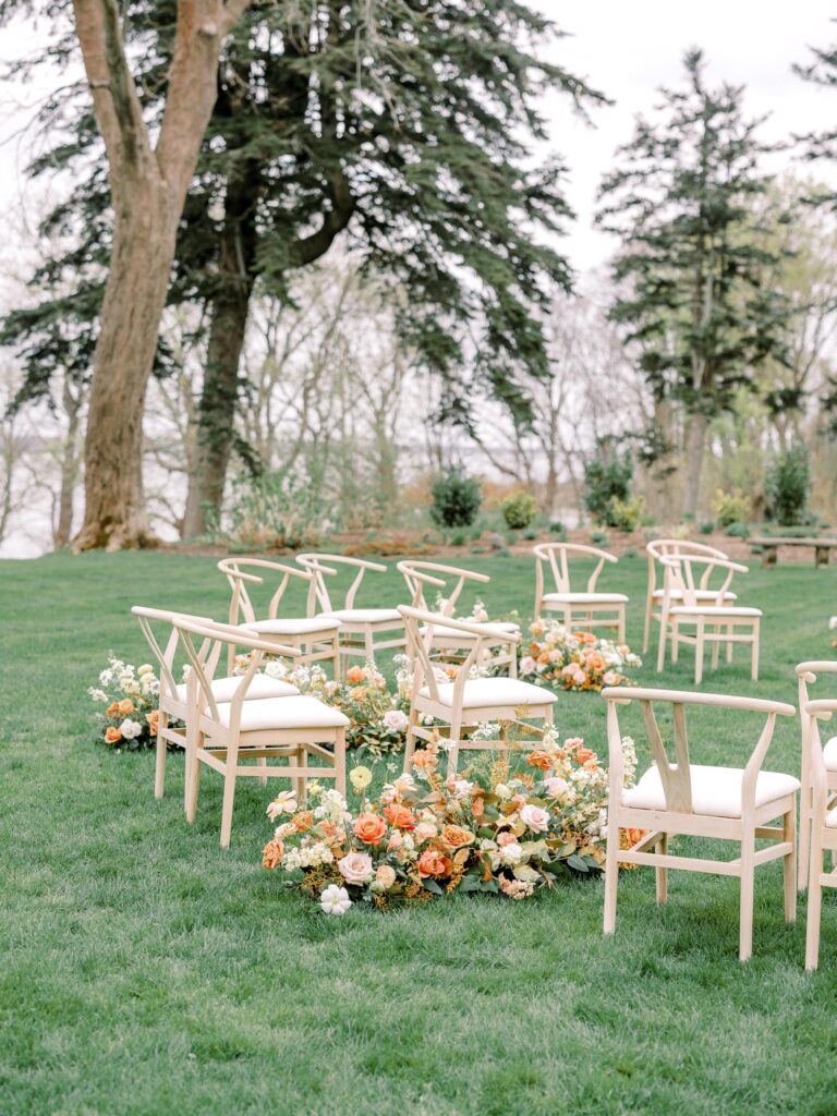Modern outdoor wedding ceremony setup with light wood chairs and lush floral aisle arrangements